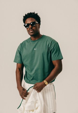 Man wearing dark sunglasses, green t-shirt, gold watch, and striped white pants, holding a white jacket against a plain background.