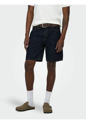 Muž oblečený v tmavých rifľových kraťasoch, bielych ponožkách, hnedých slip-on topánkach, bielej košeli a hnedom opasku, stojaci na jednoduchom pozadí.