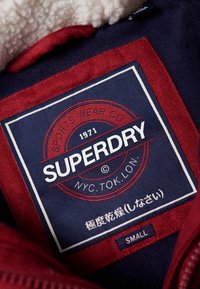 Czerwona i granatowa etykieta z polaru z białym tekstem, zawierająca nazwę marki "SUPERDRY", rok "1971" oraz instrukcje pielęgnacji. Metka z rozmiarem oznaczona jest jako "MAŁY".
