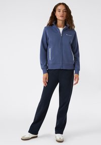 Giacca blu con zip e tasche laterali, abbinata a pantaloni blu scuro. Entrambi realizzati in tessuto morbido, adatti per un abbigliamento casual. Sneakers bianche completano il look.