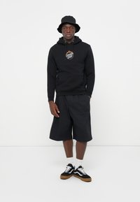 Zwarte hoodie met een logo, gecombineerd met zwarte shorts. Het model draagt een zwarte bucket hat en zwart-witte sneakers met een gumzool.