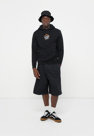 Zwarte hoodie met een logo, gecombineerd met zwarte shorts. Het model draagt een zwarte bucket hat en zwart-witte sneakers met een gumzool.