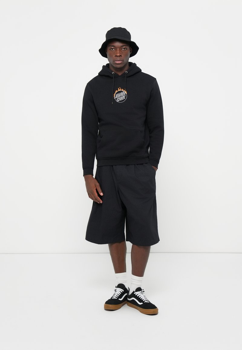 Zwarte hoodie met een logo, gecombineerd met zwarte shorts. Het model draagt een zwarte bucket hat en zwart-witte sneakers met een gumzool.