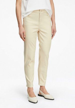 Kobieta ubrana w beżowe, przylegające spodnie z wysokim stanem oraz białe buty na obcasie ze zapięciem z tyłu, stojąca na tle białego, jednolitego tła.