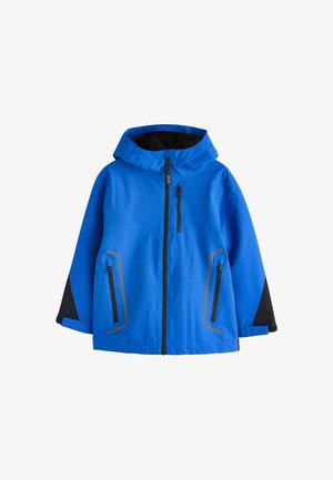 Jongen met krullend haar die een blauwe jas draagt met zwarte details en een wit hemd, kijkt iets naar de zijkant tegen een effen achtergrond.