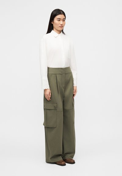 Chemise blanche à col, pantalon cargo vert avec poches latérales, et chaussures marron sans lacets. Le tissu semble lisse ; la coupe est ample.