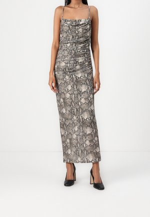 Femme portant une robe sans manches, longue jusqu'aux chevilles, imprimé serpent avec des fines bretelles et des chaussures à talons hauts noires, debout devant un fond blanc uni.