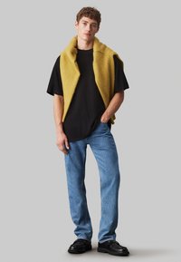 Gilet giallo, texturizzato, senza maniche sopra una maglietta nera. Jeans blu a taglio dritto e scarpe nere spesse. Sfondo grigio neutro.