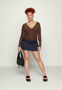 Blusa castanha de mangas longas com decote em V, combinada com uma saia mini azul marinho com riscas. A modelo segura uma mala preta e usa sandálias de plataforma pretas.