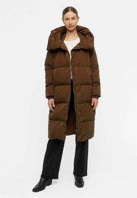 Cappotto imbottito marrone con colletto alto, grande cappuccio, design trapuntato, lunghezza fino al ginocchio, chiusura frontale con zip e bottoni a pressione. Abbinato a pantaloni neri.
