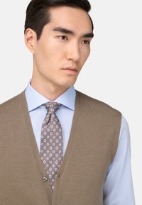 Béžový pletený sveter s výstrihom do V, nosený cez svetlomodrú košeľu. Sivá kravata so vzorom kvetov v modrých odtieňoch. Hladká textúra.