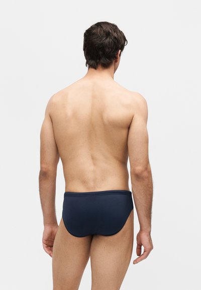 Άνδρας με κοντά σκούρα μαλλιά, γυμνή πλάτη, φοράει σκούρο μπλε μαγιό, στέκεται μπροστά σε ένα απλό ανοιχτό γκρι φόντο, με την πλάτη προς την κάμερα.