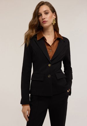 Veste noire sur mesure avec des boutons dorés, comportant deux poches avant et une coupe ajustée. Portée par-dessus une chemise marron à col.