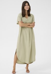 Világoszöld, térd fölé érő ruha V-nyakkivágással, rövid ujjakkal, oldalhasítékokkal és laza szabással, puha anyagból készült.