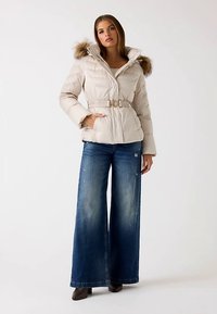 Bež prešita jakna s kapuco in umetnim krznom ob robu, zožen pas s pasom, kombinirana s širokimi kavbojkami in rjavimi gležnjarji.