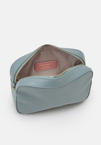 Jasnoniebieska skórzana kosmetyczka z złotym zamkiem, o zaokrąglonym kształcie i beżowym wnętrzu. Posiada czerwoną etykietę z napisem "Autentyczna włoska skóra".