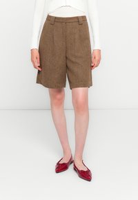 Frau, die braune Heringbone-Knie-Shorts, ein weißes gestreiftes Langarmoberteil und rote spitze Ballettschuhe trägt, steht vor einem schlichten Hintergrund.
