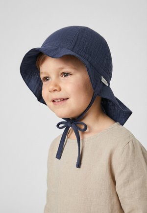 Young child wearing a navy blue sun hat with chin straps tied in a bow and a beige long-sleeve shirt, looking slightly to the side.