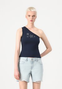 En-skulder marineblå tanktop med stjernemønster, som har en tettsittende silhuett og glatt bomullsstoff, sammen med lyse denimshorts.