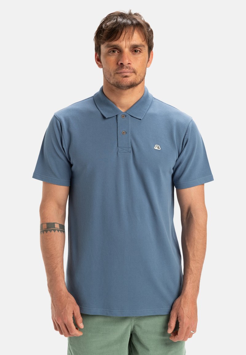 Blå bomullspolo med treknappsknäppning, kortärmade och en liten logotyp på vänster bröst. Platt stickad krage, avslappnad passform.