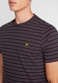 Camiseta de manga corta a rayas negras, rojas y blancas, hecha de algodón, con un logo bordado amarillo en el área del pecho izquierdo.