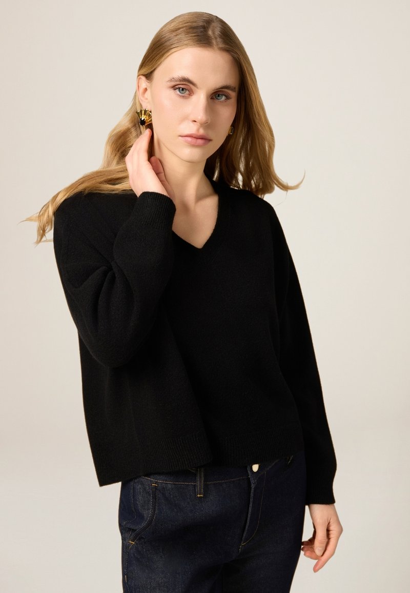 Schwarzer V-Ausschnitt-Pullover aus weichem, strukturiertem Stoff, mit überschnittenen Schultern und gerippten Bündchen. Kombiniert mit dunkler Jeans.