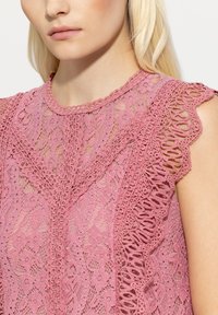 Das pinkfarbene Spitzenoberteil zeichnet sich durch aufwendige florale Muster mit gewellten Rändern aus. Der leichte Stoff hat eine strukturierte Oberfläche und einen runden Ausschnitt.