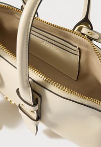 Krém bőr kézitáska arany színű fémalkatrészekkel, belső cipzáras zsebbel, sima textúrával és varrással díszített szegélyekkel.