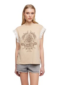 Donna che indossa un top beige senza maniche con maniche a frange e un motivo floreale e di uccelli marrone, abbinato a pantaloni corti in denim grigio chiaro.