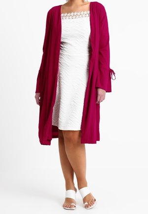 Woman wearing a white textured knee-length dress with floral lace neckline and a long magenta open-front cardigan.