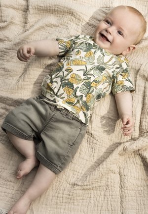 Chemise à manches courtes avec un motif de feuilles tropicales et d'ananas en vert et jaune, accompagnée de shorts en coton gris, posée sur une couverture texturée beige.