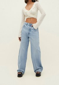 Top blanco de encaje de manga larga en envoltura combinado con jeans de pierna ancha de cintura alta en azul claro y zapatos de plataforma negros gruesos. Fondo claro.