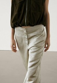 Personne portant un pantalon taille haute beige et un haut sans manches à zip vert olive foncé, se tenant avec les bras relâchés le long du corps.