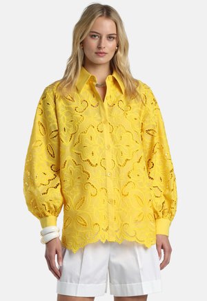 Frau trägt eine hellgelbe, florale Spitzenbluse mit Knöpfen und Puffärmeln sowie weiße Shorts, dazu weiße Armreifen als Accessoires.