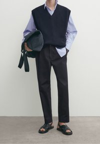 Marinblå stickad väst över en ljusblå randig skjorta, kombinerat med mörka byxor och svarta sandaler. Håller en mörk väska. Minimalistisk design.