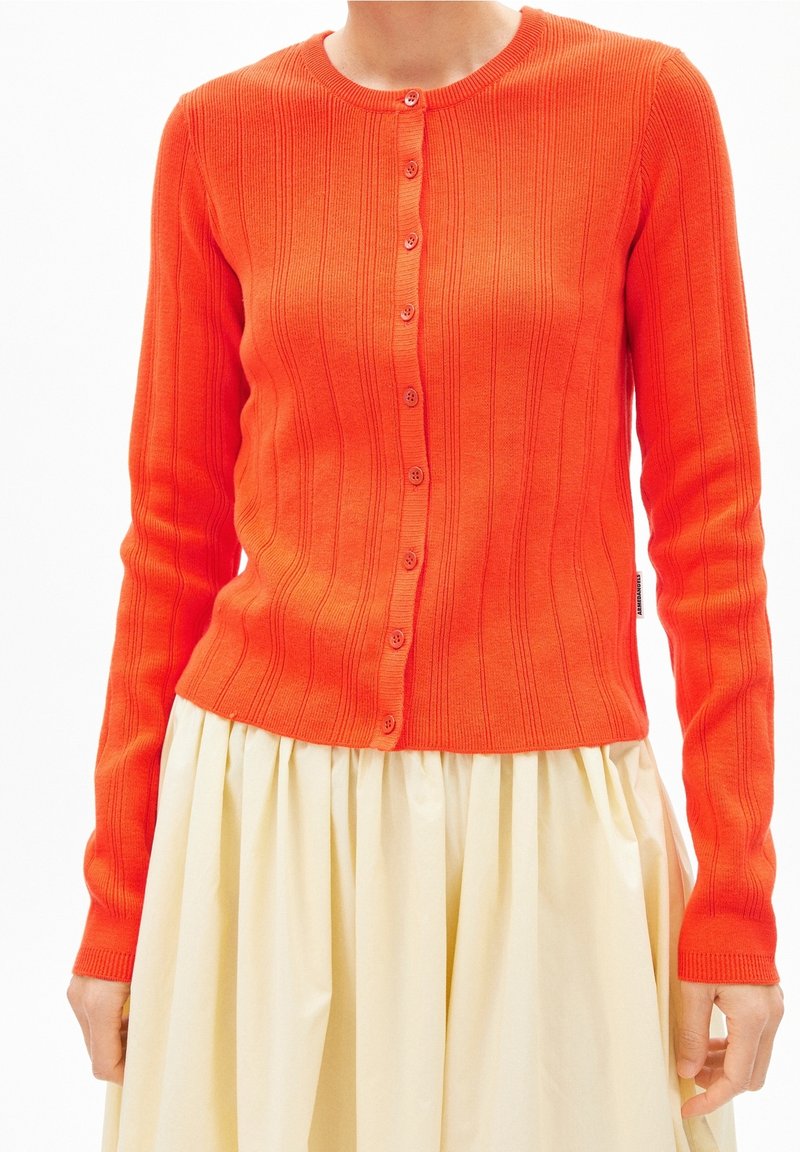 Femme portant un cardigan boutonné côtelé orange vif sur une jupe plissée crème, debout devant un fond blanc.