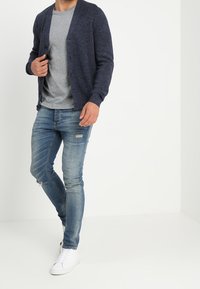 Námořnicky modrý pletený cardigan přes šedé tričko, doplněné světle modrými roztrhanými džíny a bílými teniskami. Ležérní styl s volnějším střihem.