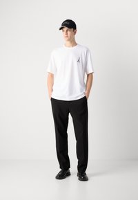 Weißes Baumwoll-T-Shirt mit kleinem schwarzem Logo, kombiniert mit schwarzen Hosen und einer schwarzen Mütze mit weißem Logo und kontrastierendem Schirm.