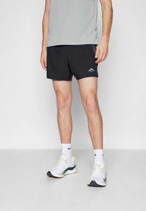 Homme portant un short de sport noir, un t-shirt gris, des chaussettes blanches Nike et des chaussures de course blanches avec des accents verts, debout sur un sol blanc.