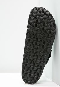 La semelle en caoutchouc noir présente un motif ondulé pour une meilleure traction. Une partie de la tige de la chaussure est visible, avec un détail de bride sur le côté.