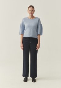Femme debout portant un pull léger à manches courtes bleu clair, un pantalon noir à jambes larges et des chaussures noires, sur un fond neutre uni.
