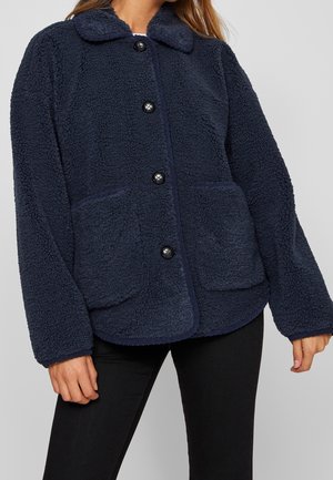 Femme portant une veste polaire bleu marine avec poches avant et boutons noirs, associée à un pantalon noir.