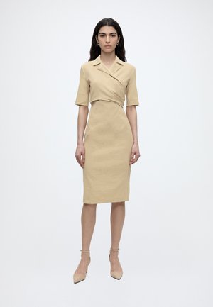 Tummahiuksinen nainen, jolla on beige polvipituinen kietaisumekko lyhyillä hihoilla ja beiget nilkkaremmi-korkokengät, seisoo yksiväristä valkoista taustaa vasten.