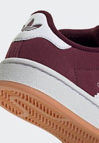 Gros plan sur le talon d'une chaussure de sport en daim bordeaux avec une semelle blanche texturée, une semelle extérieure en gomme, et un languette de talon en cuir blanc arborant le logo trèfle d'Adidas.