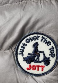 Patch rond avec la silhouette d'une personne conduisant une trottinette, texte "Just over The Top" au-dessus, et "JOTT" en rouge en dessous sur un fond en tissu.