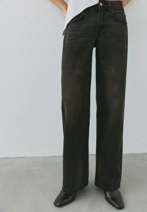 Person iført sorte jeans med vide ben og sorte spidse sko står på en grå gulv mod en hvid væg.