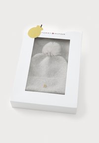 Grijze gebreide muts met een pompon bovenop, gepresenteerd in een witte doos met een doorzichtige voorkant. Inclusief details van een gouden hardware-logo.