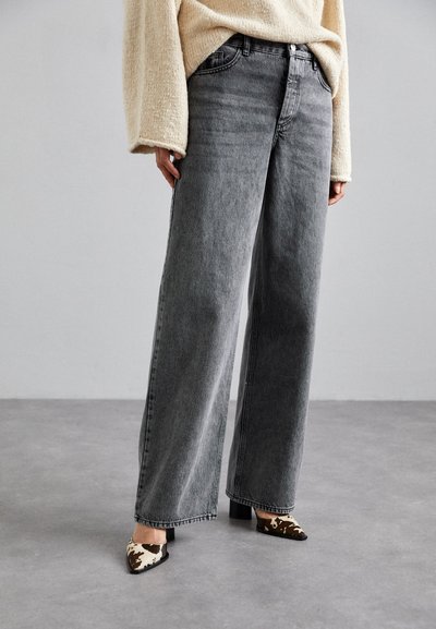Graue Jeans mit weitem Bein und hohem Bund, die ein klassisches Fünf-Taschen-Design und eine subtile, ausgebleichte Textur aufweisen, kombiniert mit gemusterten Absätzen.