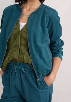 Vrouw draagt een teal corduroy jas met rits en broek, gecombineerd met een olijfgroene button-up cardigan en een wit overhemd eronder, hand in de zak.