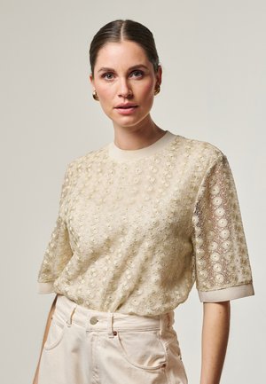 Vrouw met donker haar in een knot, die gouden oorringen draagt, een beige korte broek met bloemenkant en hoge taille crème broeken, tegen een lichte achtergrond.